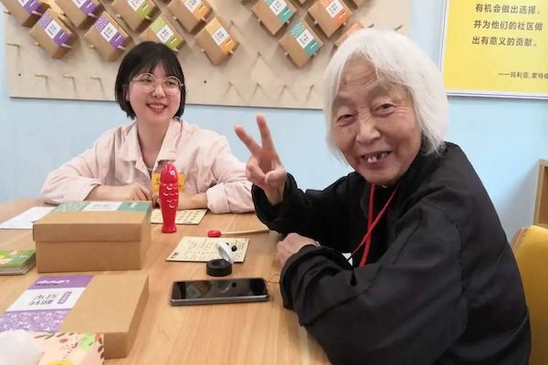 A young and elderly woman sit at a table, with the elderly woman holding a peace sign to the camera.