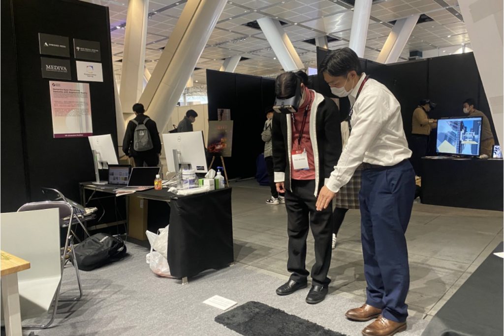 A man points to the ground as another man next to him wears a VR headset.